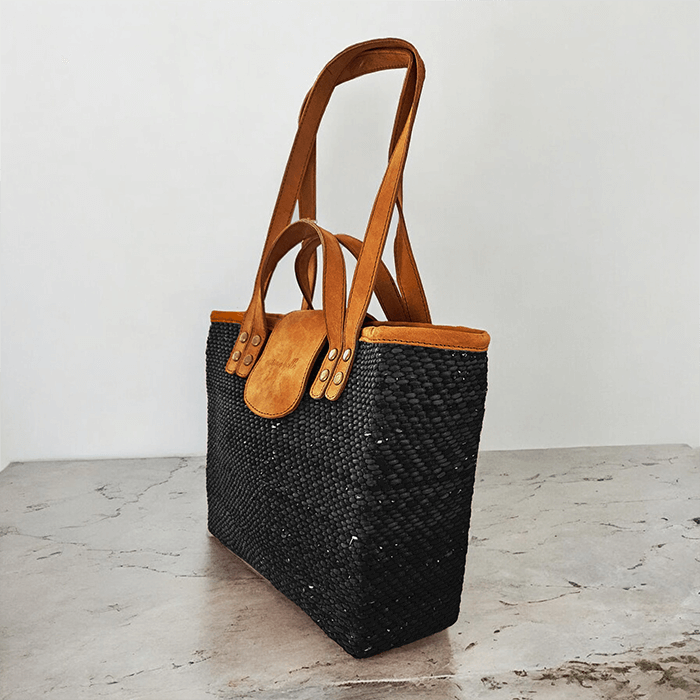 Black woven handbag with brown leather straps on a light wooden surface.