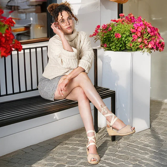 Woman sitting on a bench with flowers and a white wall in the background wearing platform espadrilles in cream