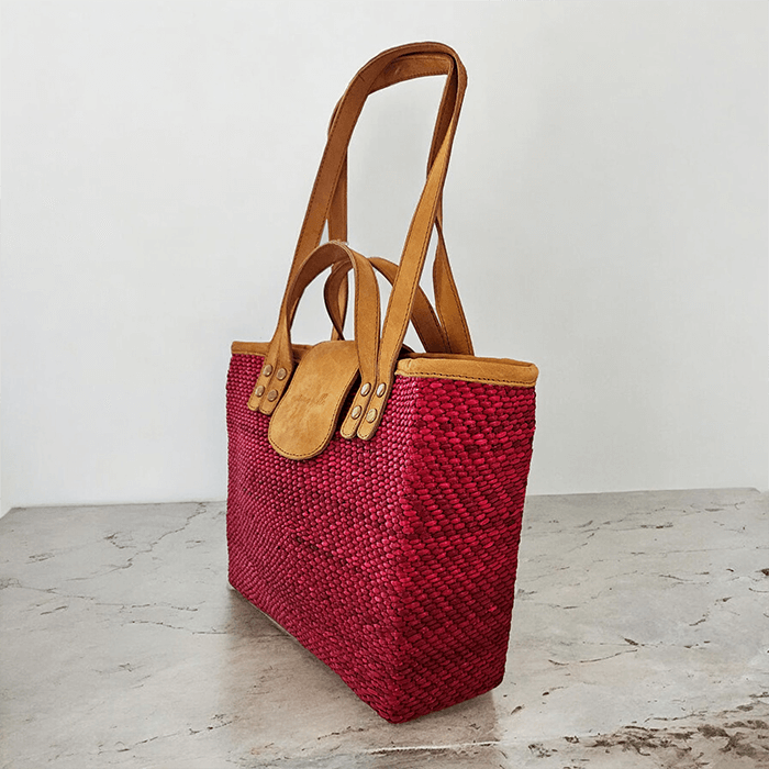 Red woven handbag with brown leather handles on a light wooden surface.