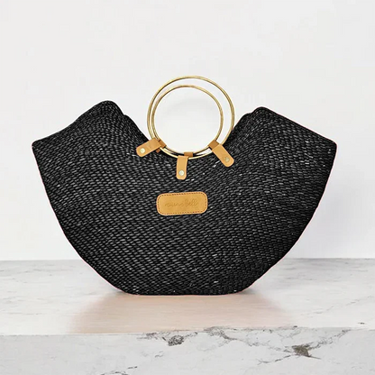 A handwoven black seagrass tote bag with brass handles, displayed against a marble background.