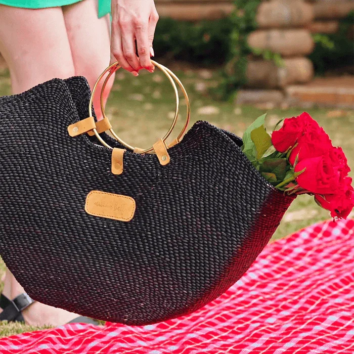 Black woven handbag with gold handles held by a person on a red and white checkered blanket.