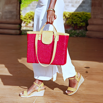 A person wearing white pants and yellow sandals is holding a vibrant red seagrass shoulder bag with brown leather handles.