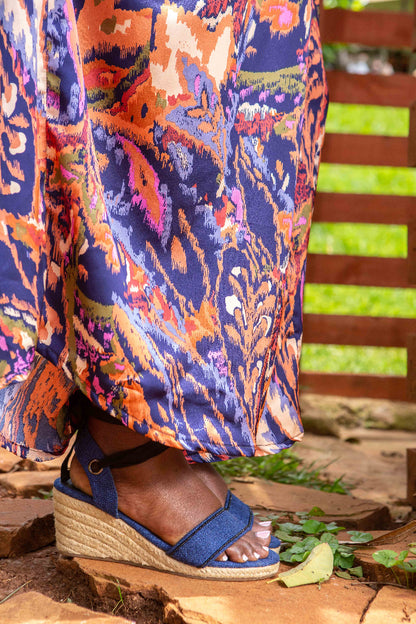 Person wearing colorful patterned pants and blue wedge sandals on a wooden floor.