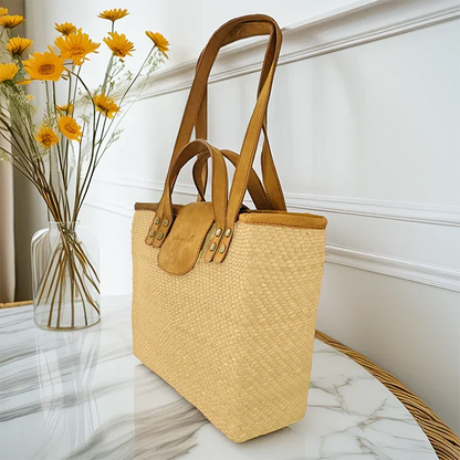 Woven tote bag with leather handles on a marble surface next to a vase of yellow flowers.