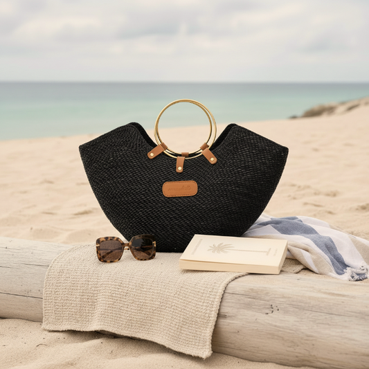 Black straw bag with gold handles on a beach with sunglasses, book, and towel.