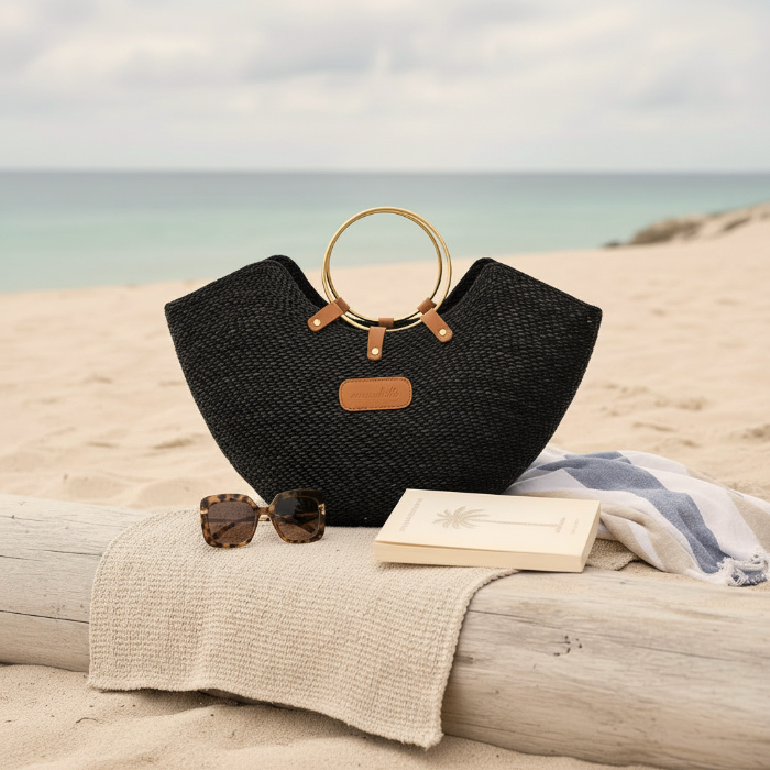 Black woven straw beach bag with brass ring handles, on a log with sunglasses and a book.