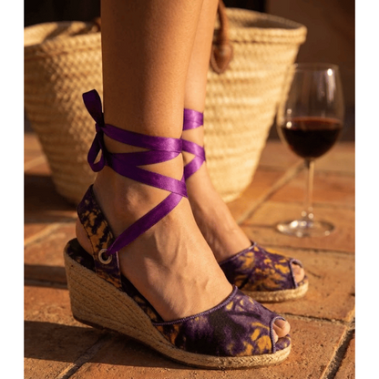 Purple wedge sandals with floral pattern worn by a person on a wooden floor.