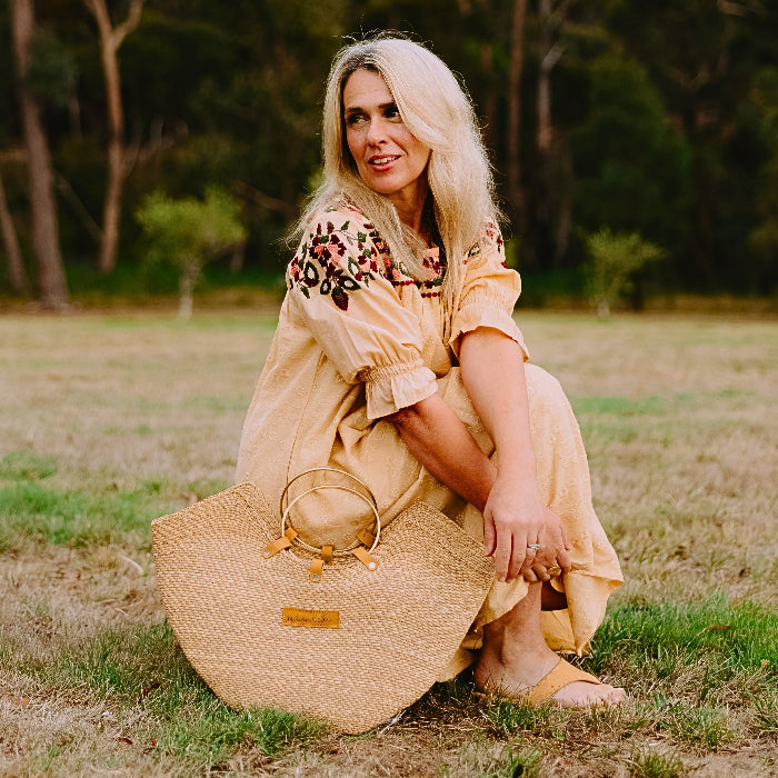 Woman in a yellow dress sitting on grass with a straw bag