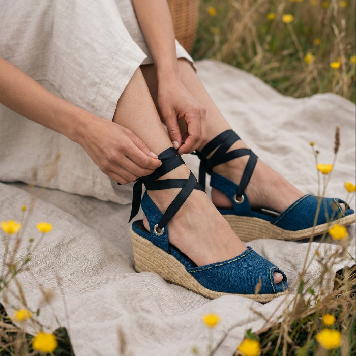 Person wearing gaia indigo wedge sandals with black straps in a natural setting with yellow flowers.