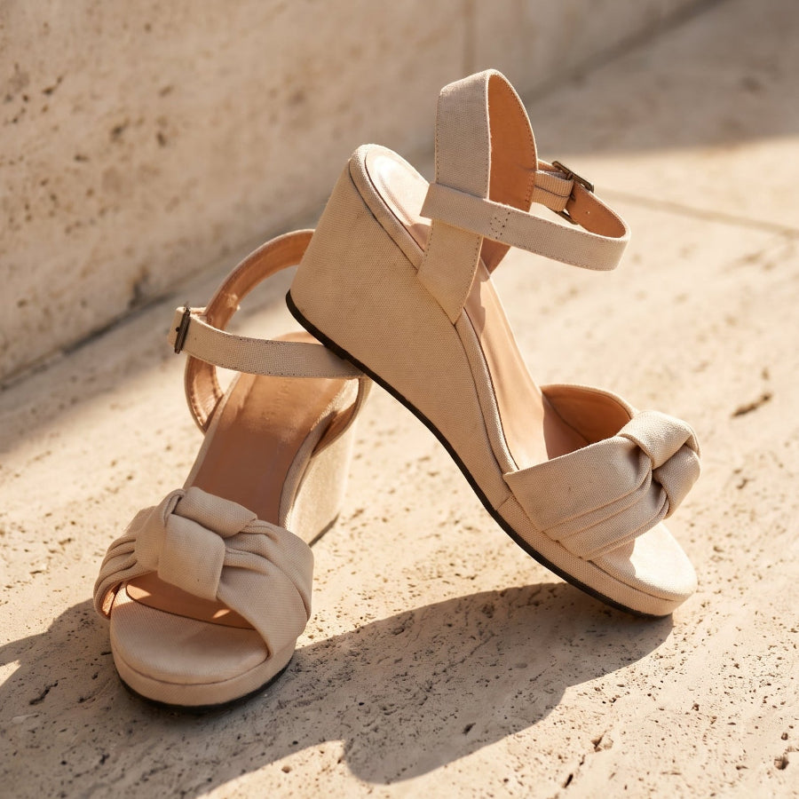 A pair of beige wedge sandals with ankle straps and a knotted front detail, photographed on a sunlit stone surface against a textured wall.