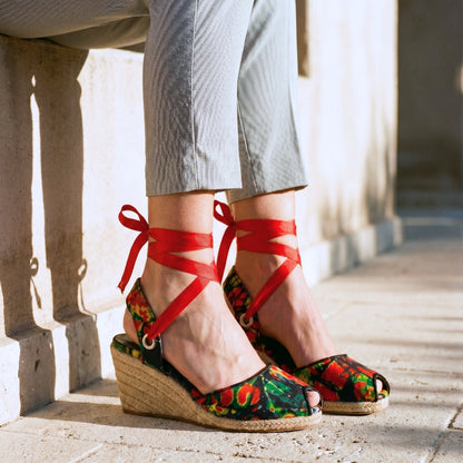 Gaia Wild Flame espadrille wedge heels with red ribbon ankle ties and printed canvas worn by a person on a stone pavement.