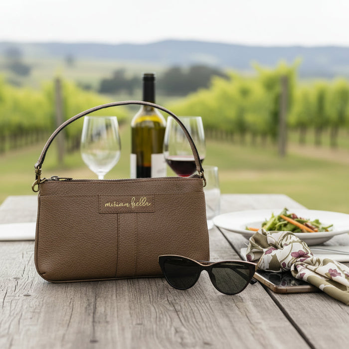 Brown handbag with 'michael kors' branding on a wooden table with wine, glasses, and food in a vineyard setting.