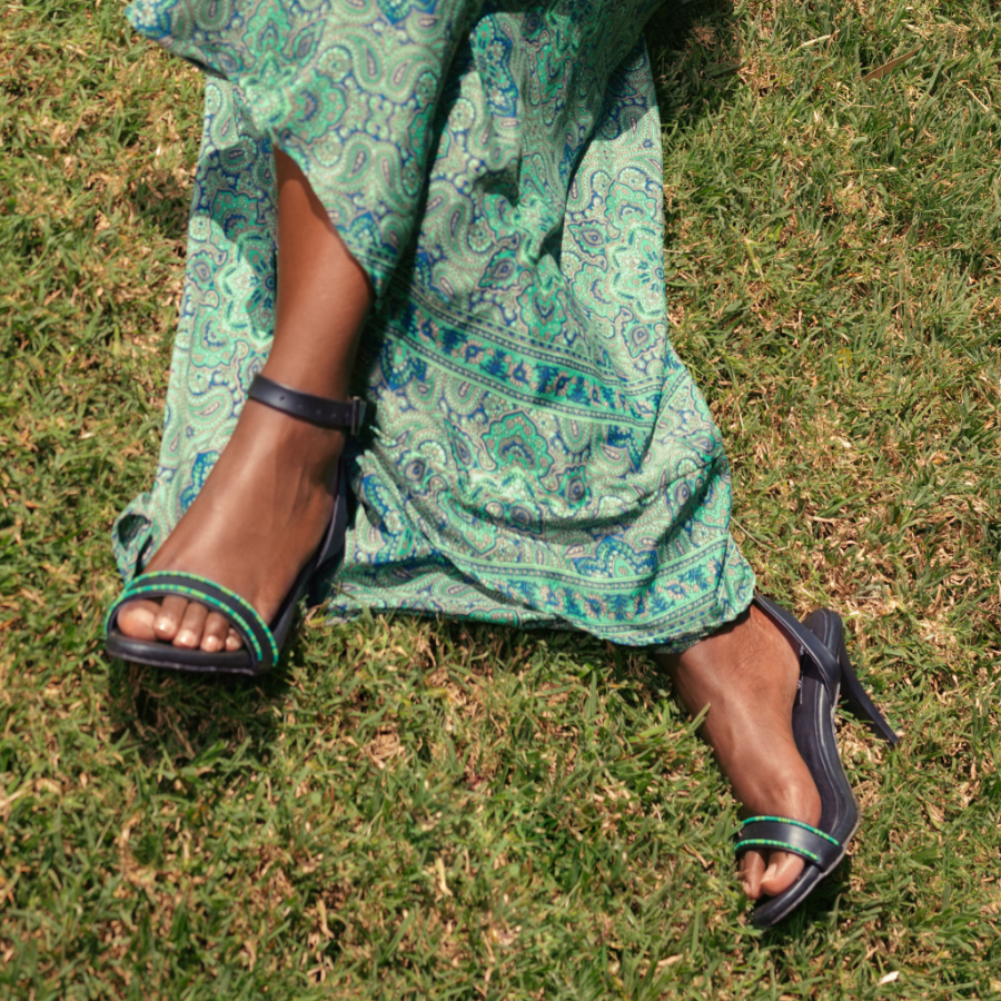 A person sitting on green grass wearing a flowing green paisley dress and black heeled sandals with green trim.