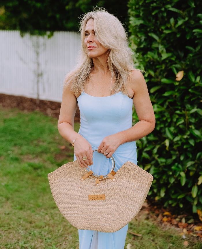 Woman in a blue dress holding a natural straw seagrass tote bag with brass handles.