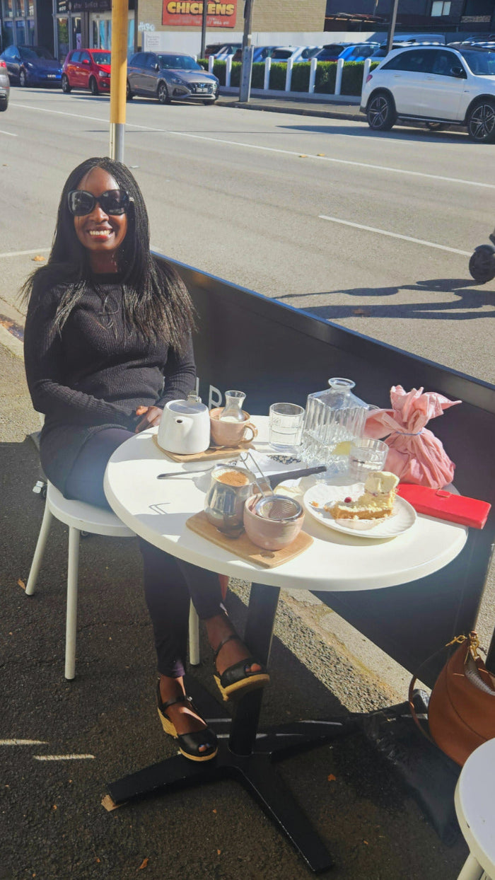 Woman sitting at an outdoor table with food and drinks, on a street corner. She is wearing black wedge espadrille sandals