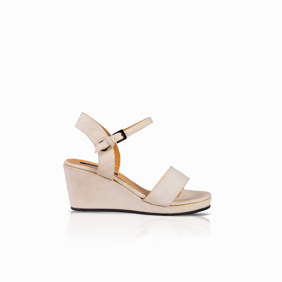 A single women's wedge sandal in a light beige or cream color, featuring a wide front strap, a slim ankle strap with a small black buckle, and a wedge heel, shown against a plain white background with a soft reflection.