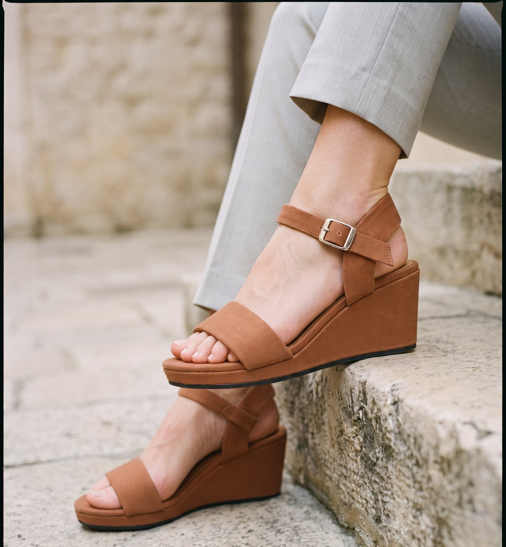A person wearing brown wedge sandals with ankle straps and a small silver buckle, seated on stone steps with light gray pants visible.