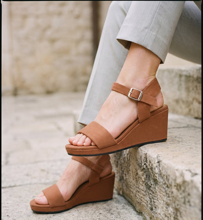 A person wearing brown wedge sandals with ankle straps and a small silver buckle, seated on stone steps with light gray pants visible.