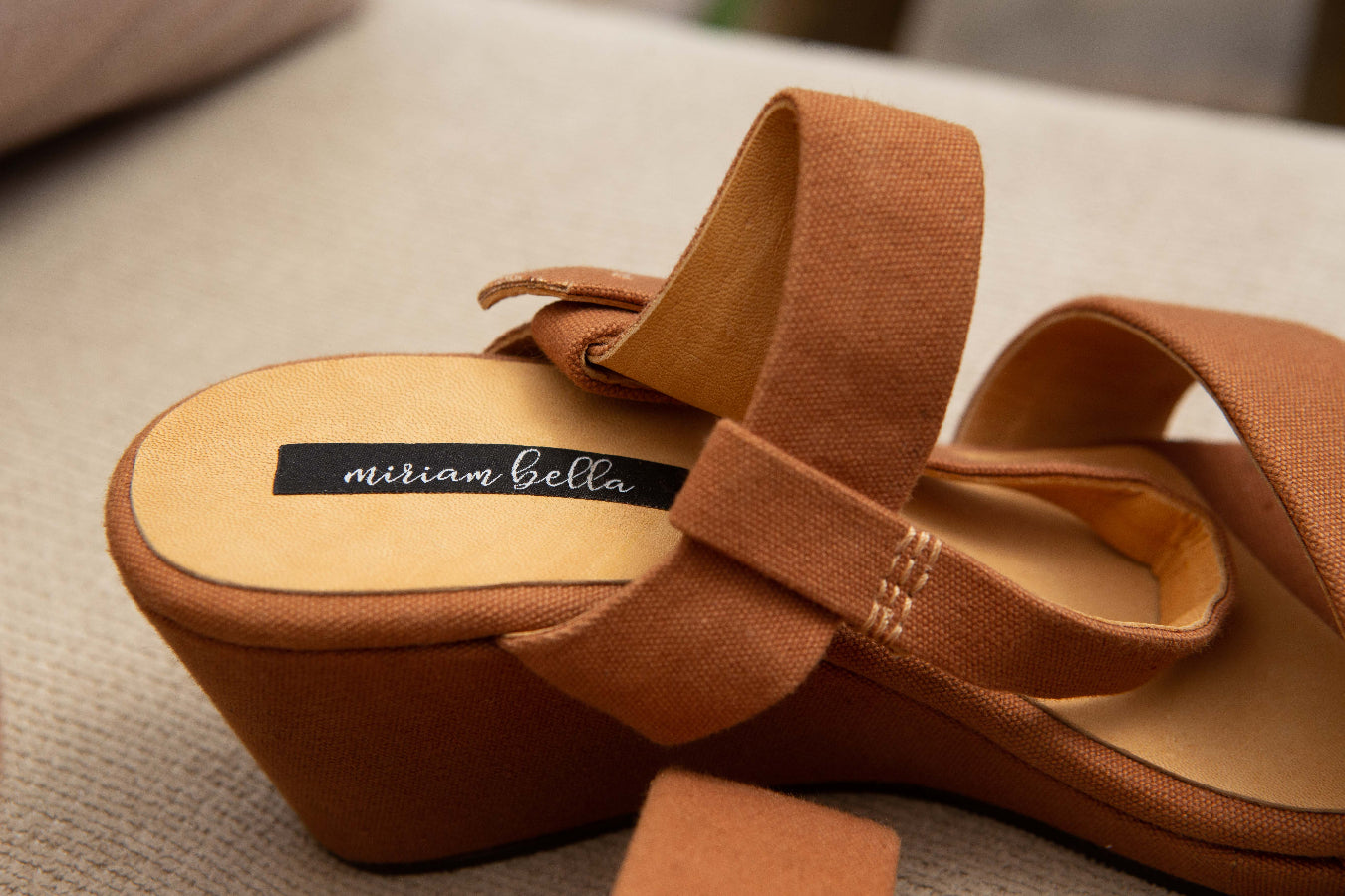 A close-up of a tan wedge sandal with wide fabric straps and a black insole label reading 'nitram bella'.