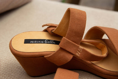 A close-up of a tan wedge sandal with wide fabric straps and a black insole label reading 'nitram bella'.