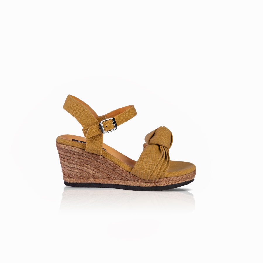 A single women's wedge sandal in mustard with a knotted detail across the instep, an adjustable ankle strap with a silver buckle, and a natural jute-wrapped wedge heel, shown from a side profile on a white background.
