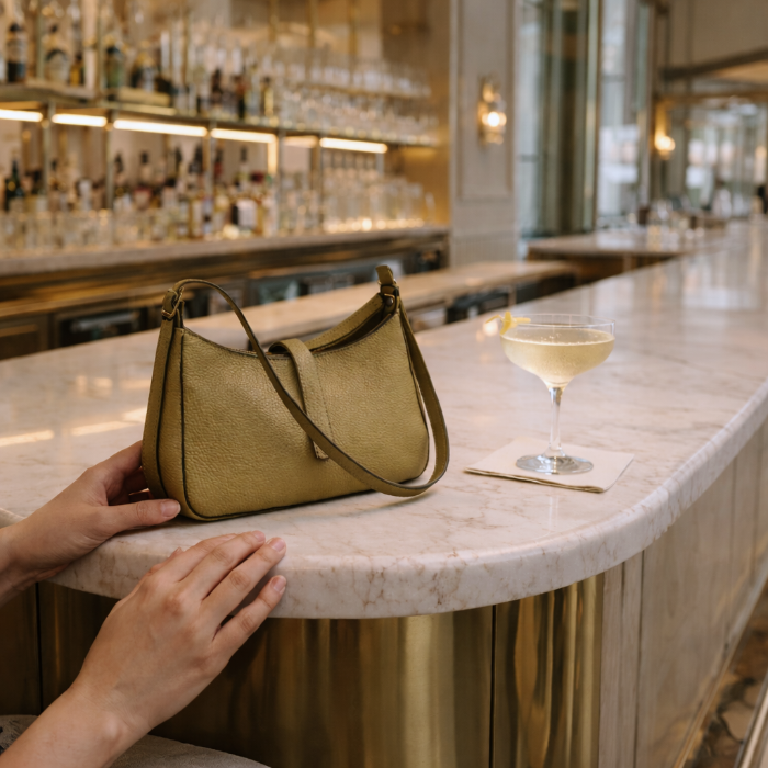 An olive leather mini shoulder handbag on a marble bar counter with a glass of white wine in the background