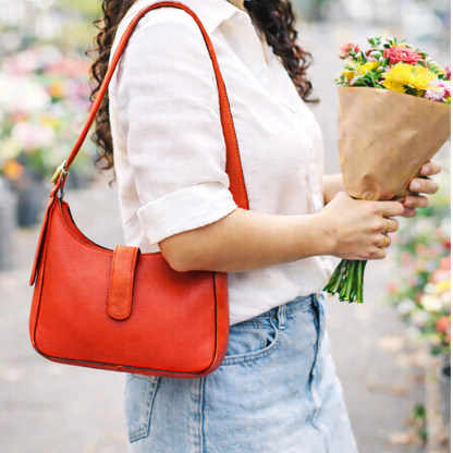Person holding a red leather shoulder bag and flowers in an outdoor setting