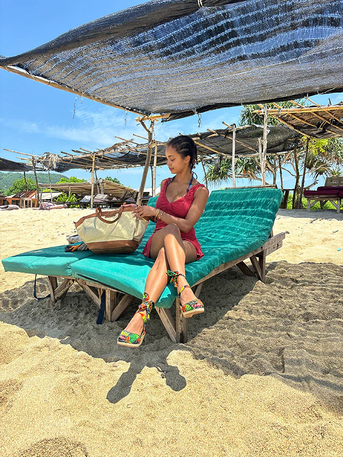 Woman in a red swimsuit and espadrille wedge sandals sitting on a green lounge chair under a thatched roof on a beach.